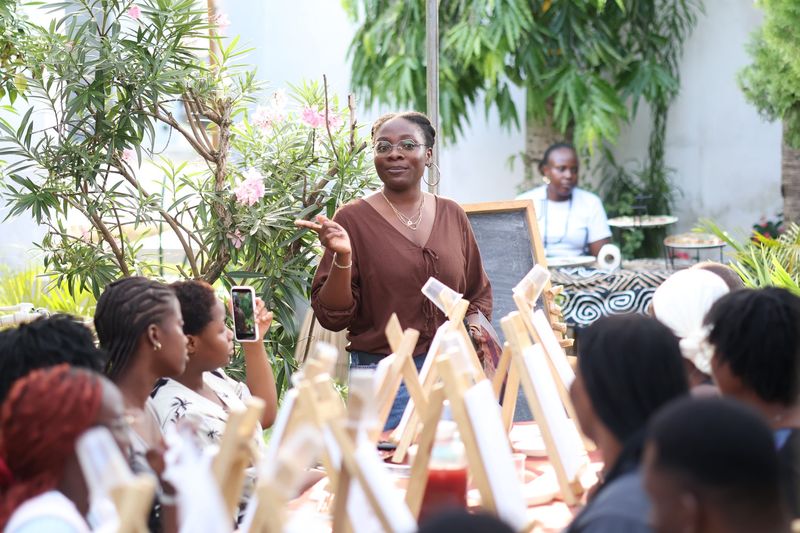 A host in brown speaks to a seated audience surrounded by painting easels at the book club