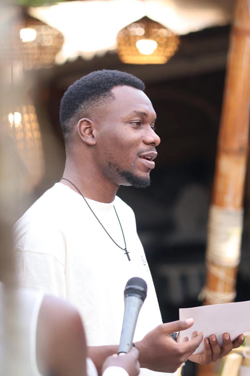 Young man speaking into a microphone and holding notes at an outdoor book club gathering lit by rattan lanterns