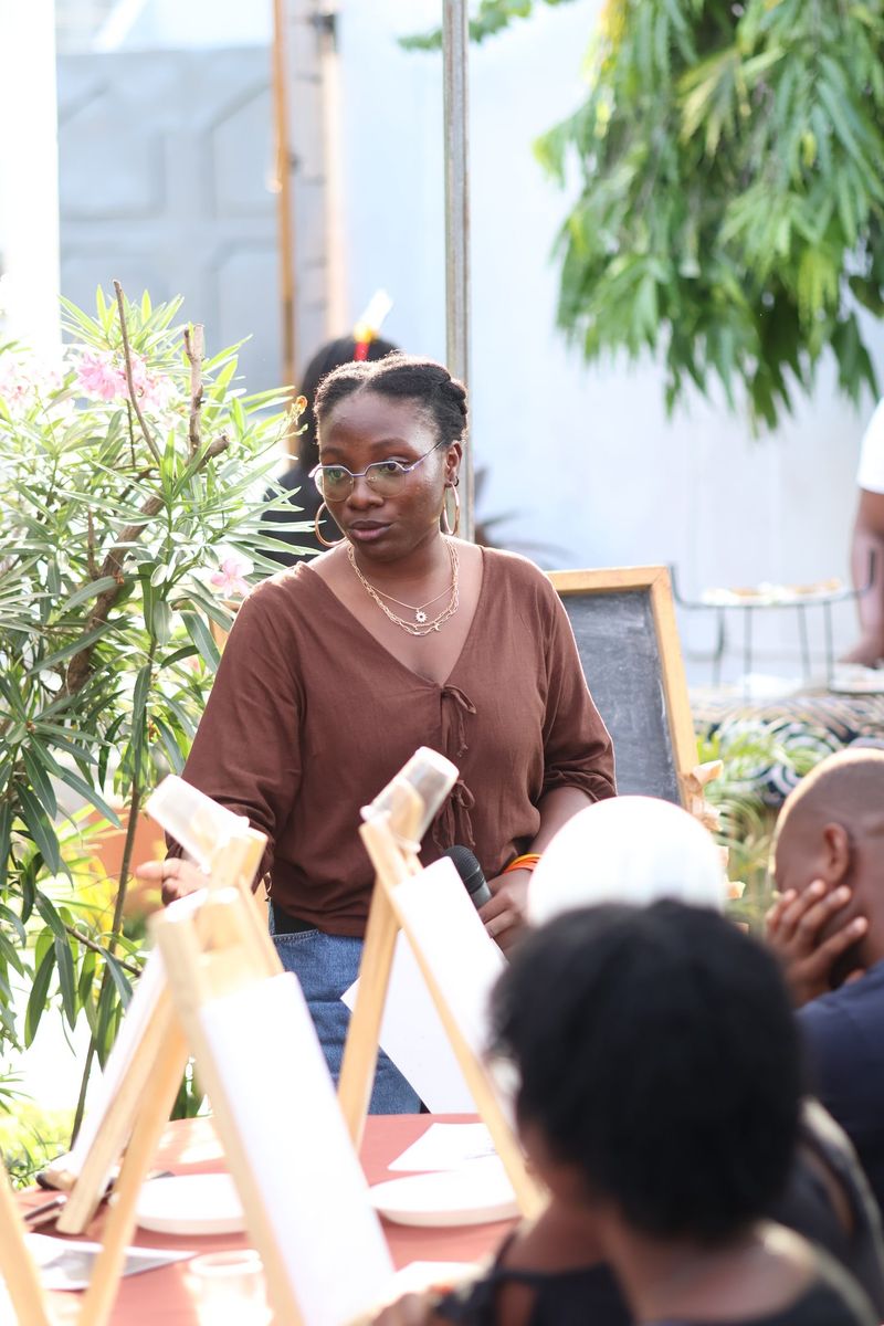 The host in brown speaks to seated guests among painting easels