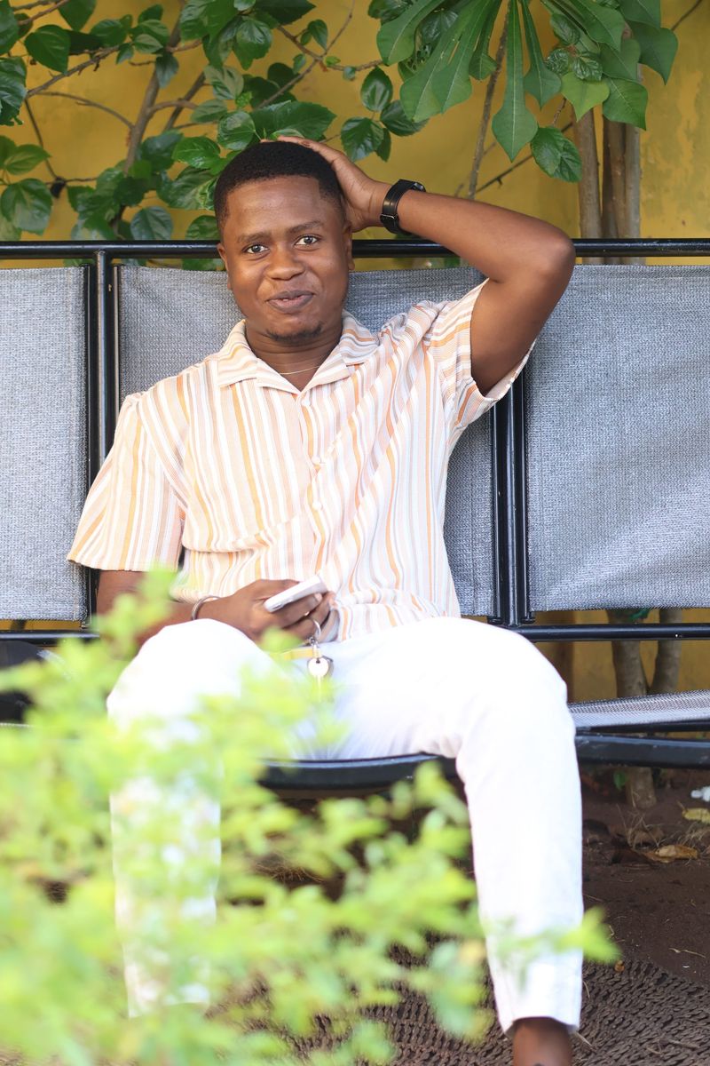 A man in a striped shirt and white trousers relaxes on a bench amid greenery