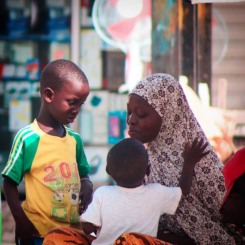 A woman in a patterned headscarf stands with two young boys on a city street, one boy in a yellow football jersey