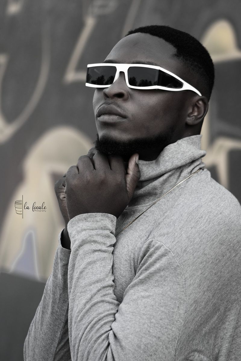A young man in white sunglasses and a grey top poses thoughtfully with a hand to his chin