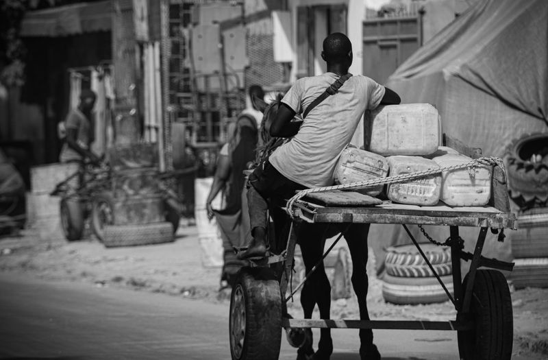 A man drives a horse-drawn cart loaded with water containers through a market street, black and white