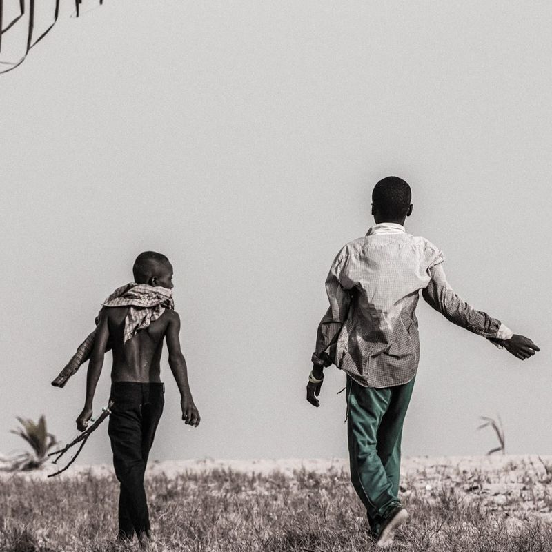 Black and white photo of two young men walking past a bare wall, one shirtless holding a slingshot