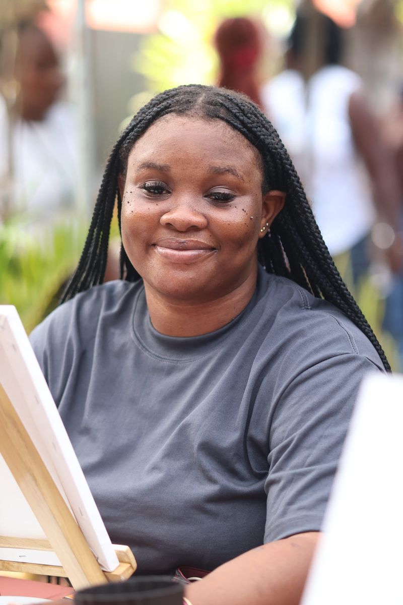 A smiling young woman with braids sits at a painting easel in a grey t-shirt
