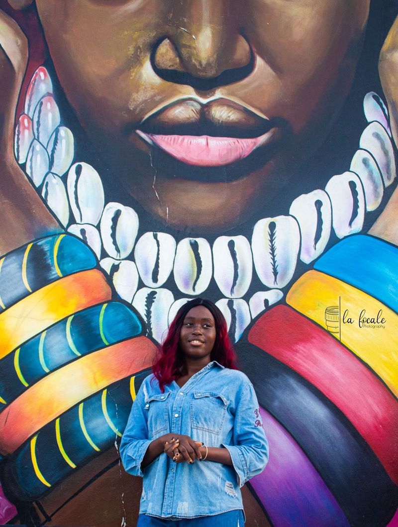 A young woman in a denim shirt stands before a vibrant mural of a face and a cowrie-shell necklace