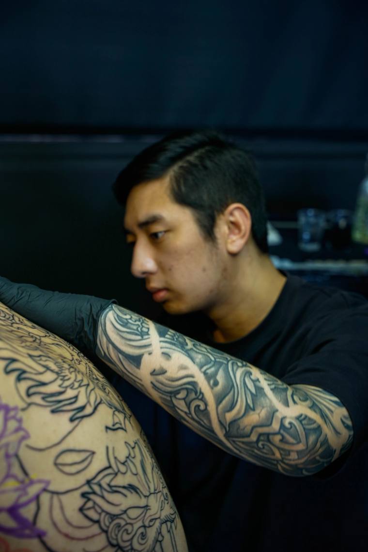 Tattoo artist in black gloves working on a large black-ink irezumi piece on a client's leg, mid-session.