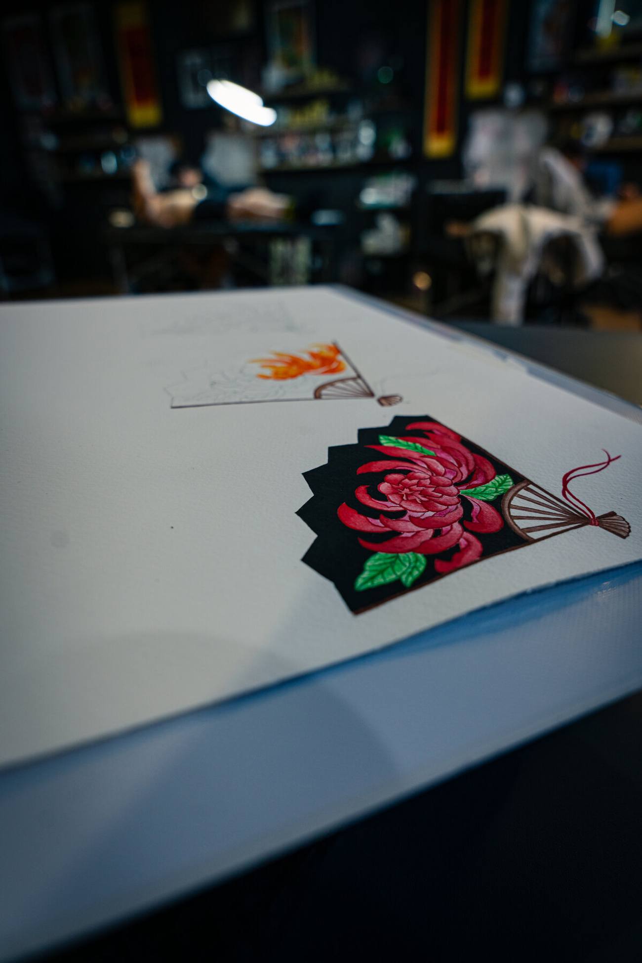 Mandy - Oriental fan flash, a crimson chrysanthemum blooms across a black folding fan, drawn in watercolour alongside a second design in progress.