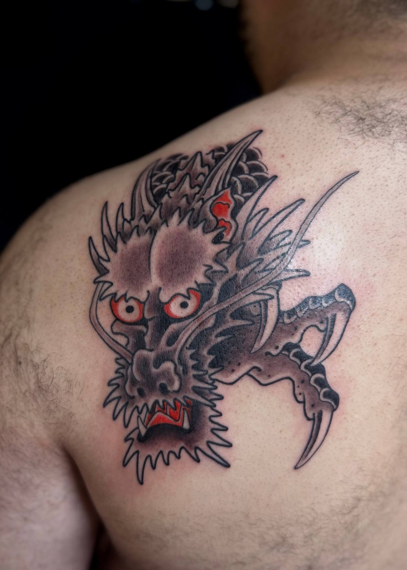 Kevin - A fierce neo-traditional dragon rendered in black, grey and red, snarling across the shoulder with detailed scales and a razor-sharp talon.