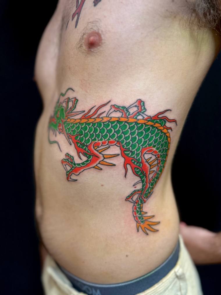 Colourful green-scaled Asian dragon with orange flames inked on a man's ribs in traditional irezumi style.