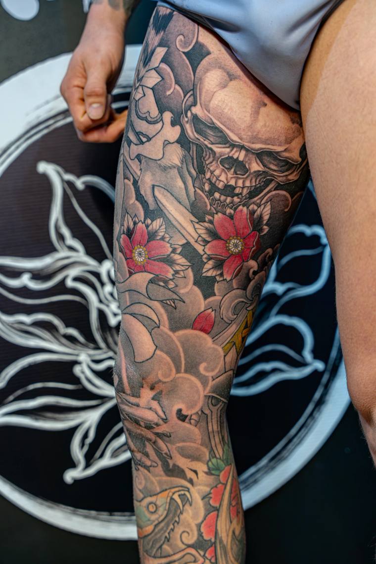 Japanese-style calf tattoo featuring a skull, red peonies, wind bars, and a dragon head in black, grey, and red ink.