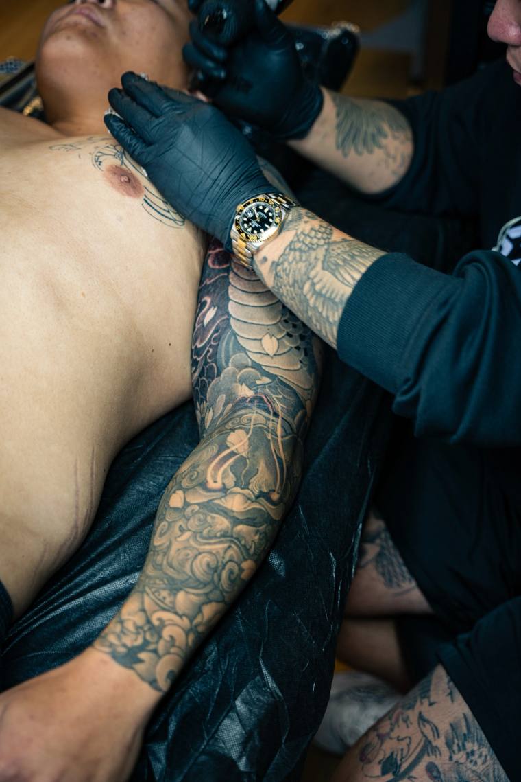 Mid-session irezumi sleeve being tattooed on a client's arm, showing koi scales, clouds and waves while the artist works with