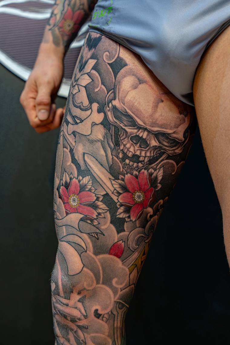 Large greywash skull tattoo surrounded by red cherry blossoms and swirling Japanese-style clouds on a thigh.
