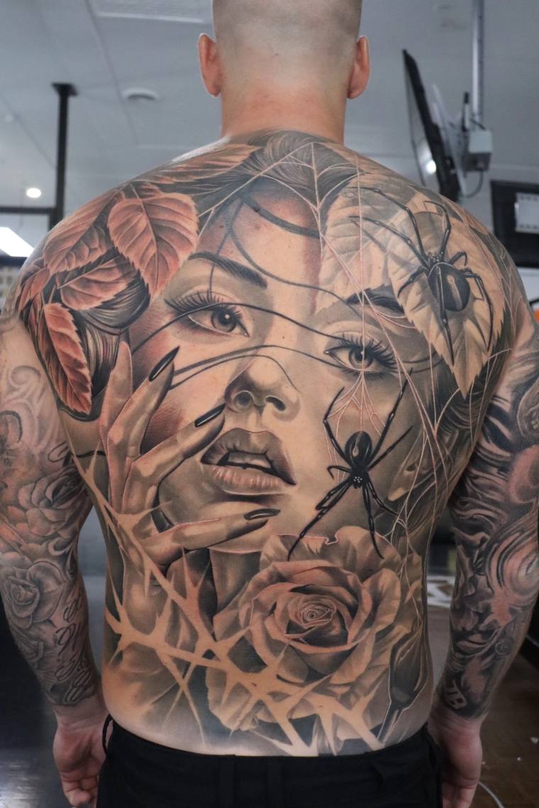 Large black and grey back tattoo featuring a woman's face surrounded by roses, leaves, a spider web, and black widow spiders.