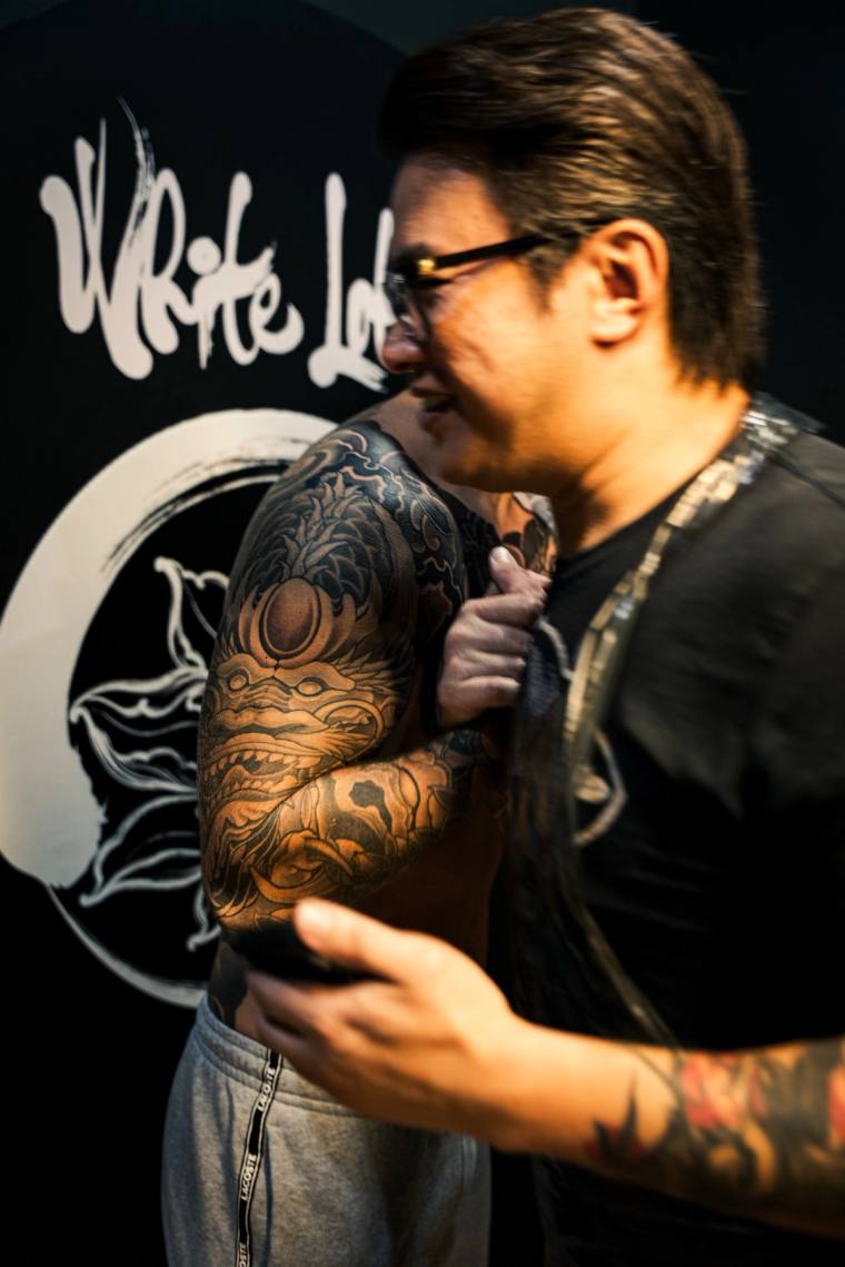 Smiling tattoo artist in glasses wearing a black t-shirt, turning to show off his finished irezumi sleeve featuring a daruma 