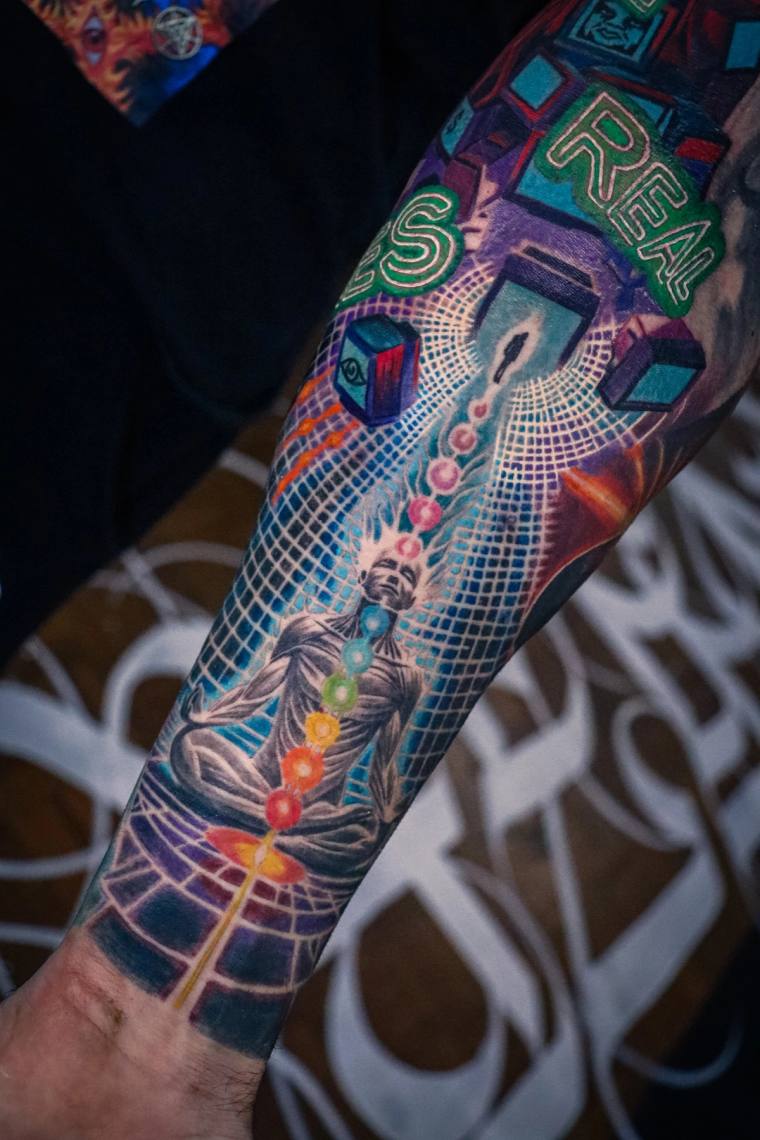 Vibrant full-sleeve forearm tattoo of a meditating Buddha with rainbow chakra points, geometric cubes, and green RELAX letter