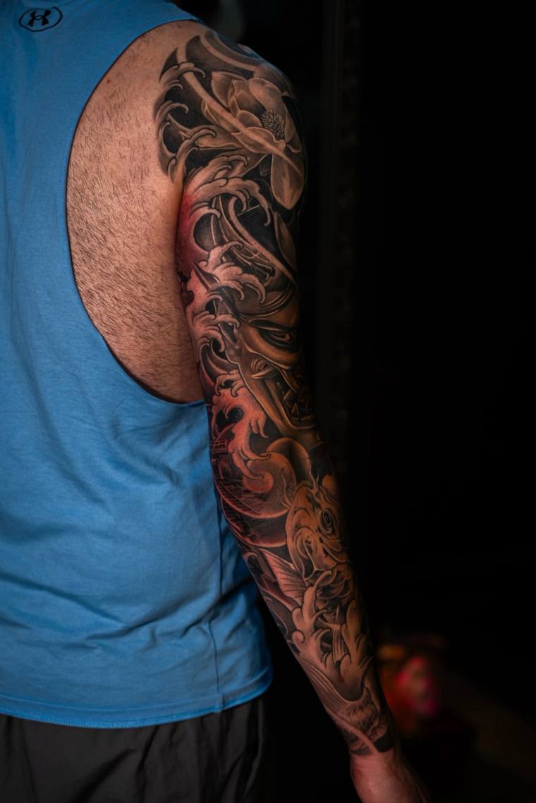 Finished black and grey full sleeve tattoo featuring a hannya mask, poppies, and flowing flame motifs on a man's left arm.