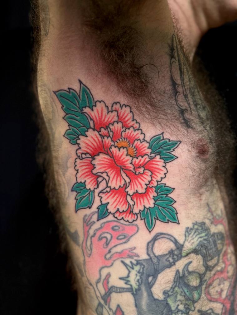 A vibrant red Japanese peony with green leaves tattooed on an arm, with a hannya mask visible below as part of a sleeve.