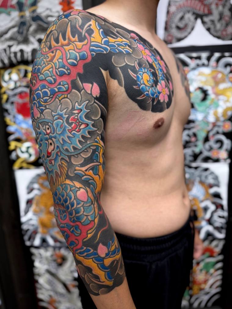 Man photographed in profile showing a vibrant Japanese-style irezumi full sleeve and chest piece featuring a dragon, koi, peo
