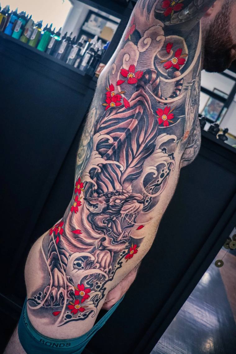 Finished irezumi full sleeve featuring a roaring tiger's head framed by ocean waves and red cherry blossoms on a man's arm.