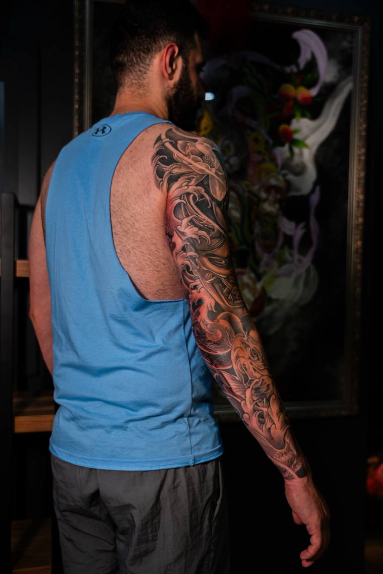 Man in light blue tank top displaying a finished Japanese-style irezumi full sleeve tattoo featuring koi and crashing waves o