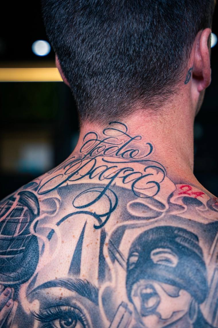 Back of a man's head and shoulders showing 'Todo Mi Vida' script on the neck and detailed black-grey portrait tattoos across 