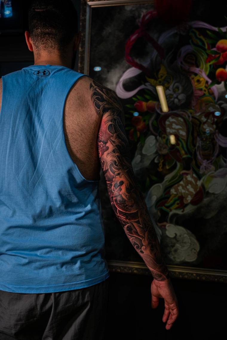 Back view of a man in a blue tank top displaying a vibrant, fully healed irezumi sleeve with waves, koi, and a Japanese deity