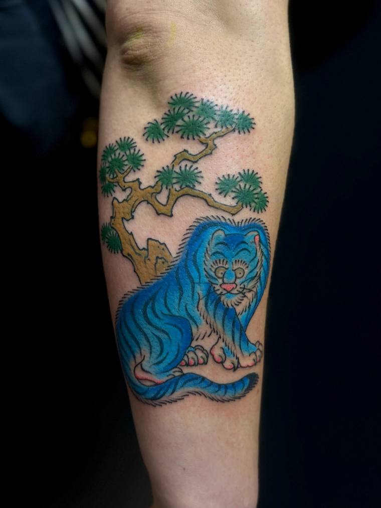 A vivid blue tiger with black stripes crouches beneath a green pine tree with a golden trunk, inked on a forearm in tradition