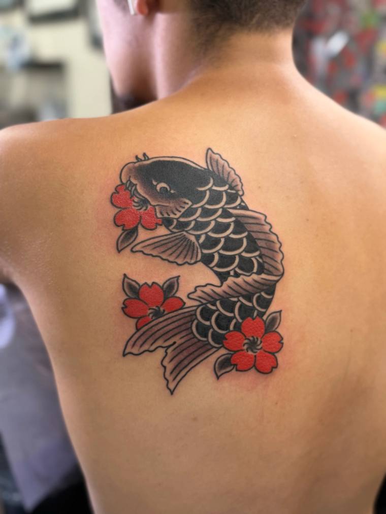 A koi fish tattoo with red cherry blossoms on a person's upper back, rendered in black ink with red floral accents.