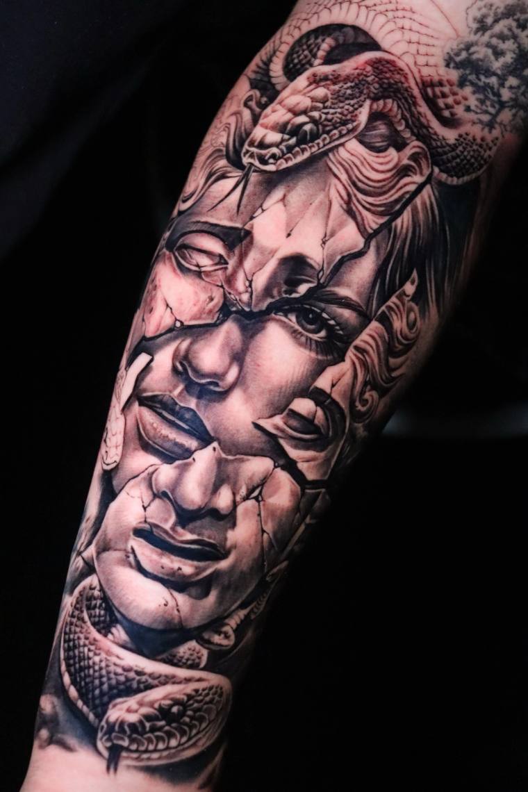 Black and grey forearm tattoo of Medusa's face rendered as cracked stone, with a coiled snake at the crown of the head.