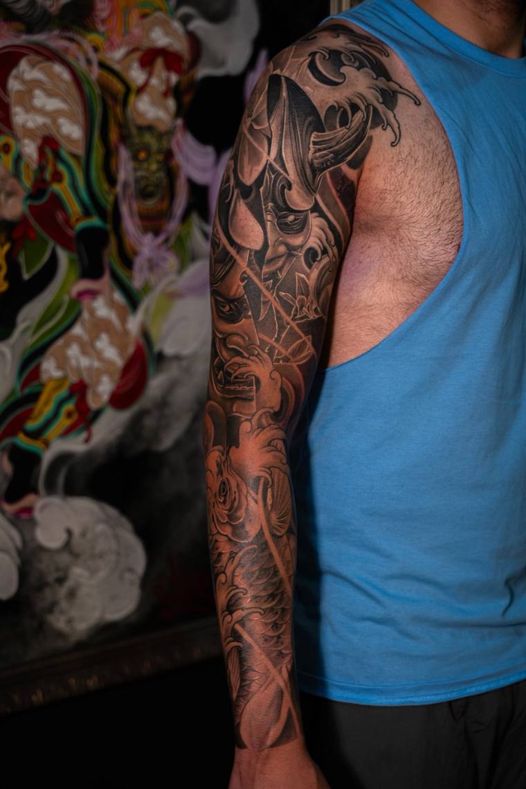 Man in blue tank top displaying a finished black and grey irezumi sleeve tattoo with a hannya mask and wave motifs on his lef