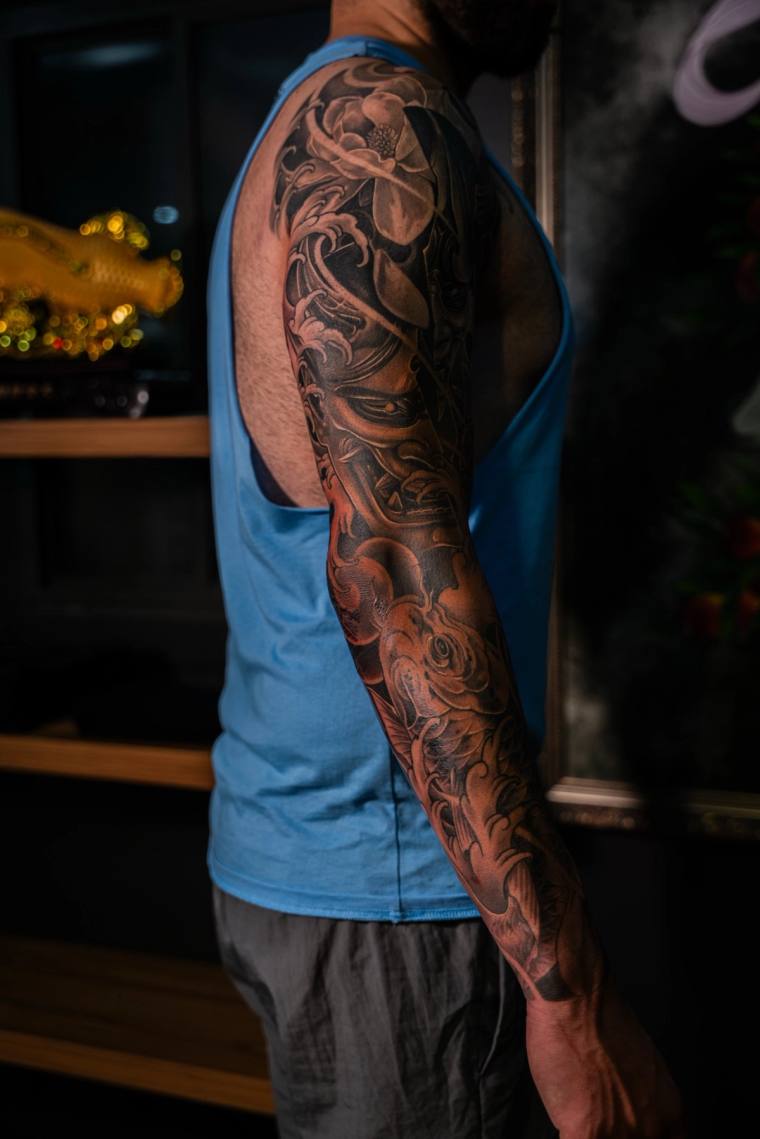Black and grey Japanese sleeve tattoo featuring a fierce hannya mask, peony blooms, and swirling waves from shoulder to wrist