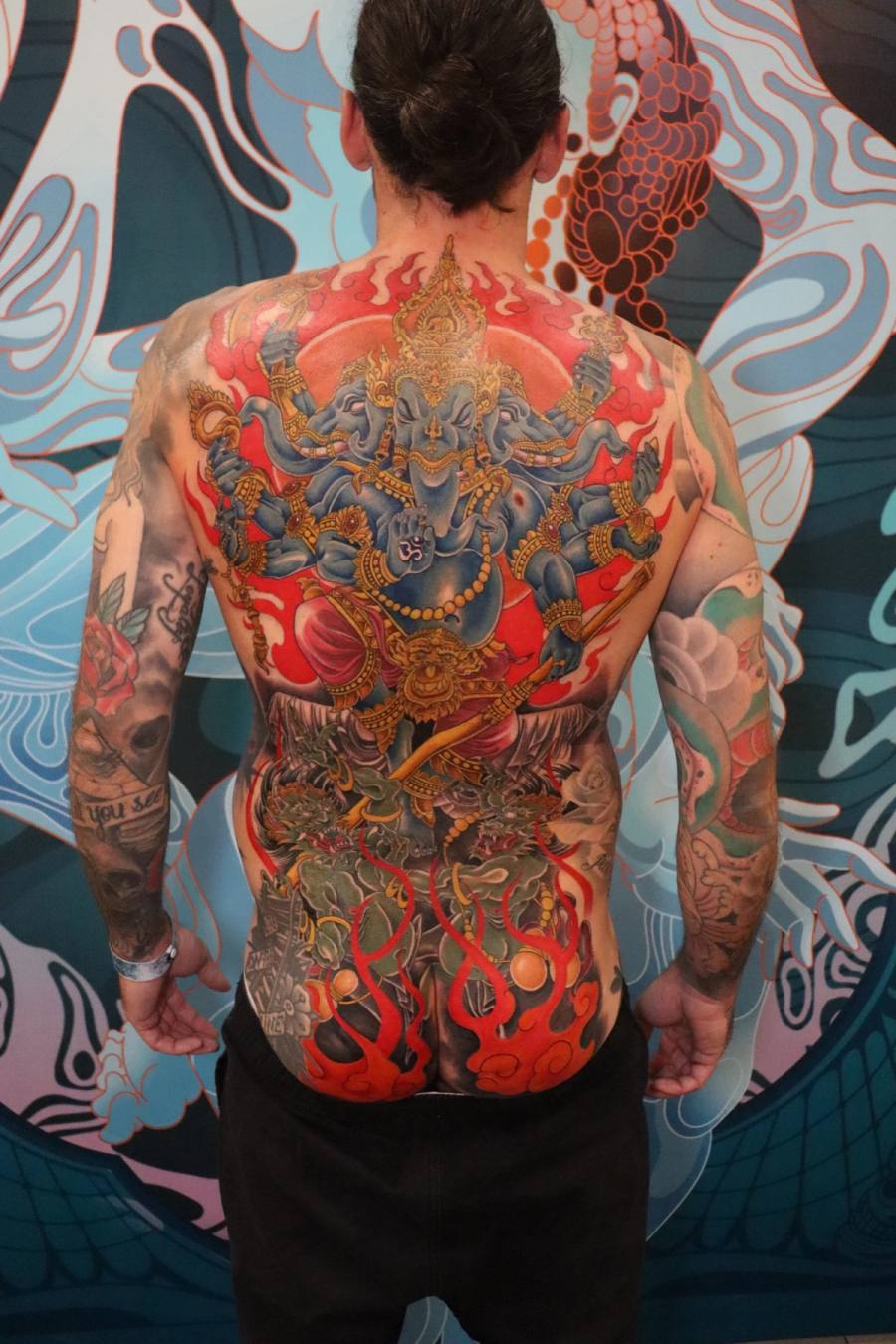 A bold full-back irezumi piece centered on a multi-armed Ganesha-like deity rising from flames and demons, paired with matching sleeves. by Rodney