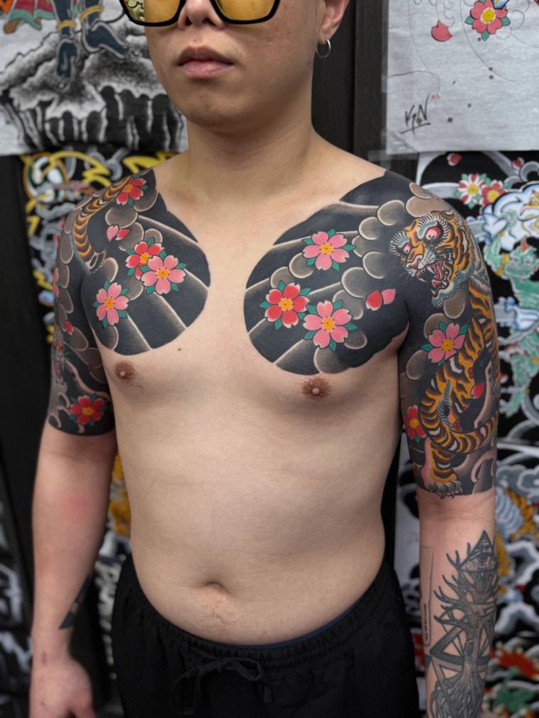 Shirtless person displaying irezumi shoulder and sleeve tattoos featuring cherry blossoms, wind bars, clouds, and a fierce ti