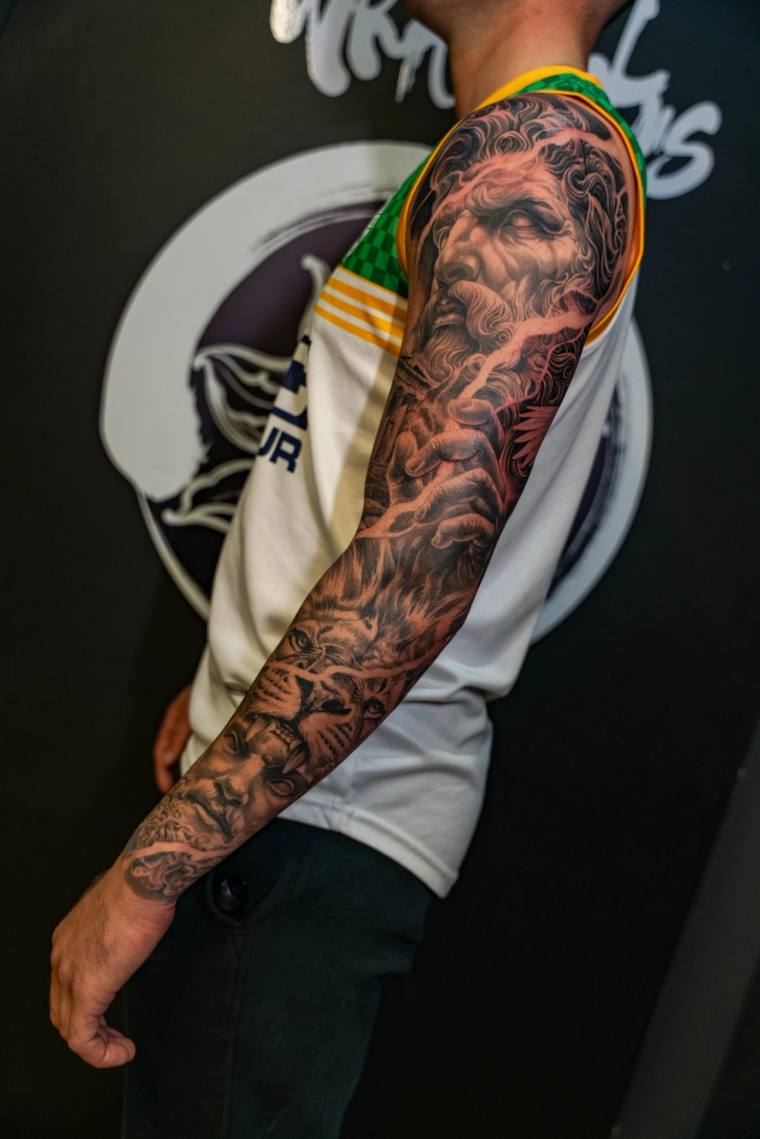 Finished black and grey full sleeve tattoo featuring a bearded Zeus figure with lightning bolts on the shoulder and a roaring