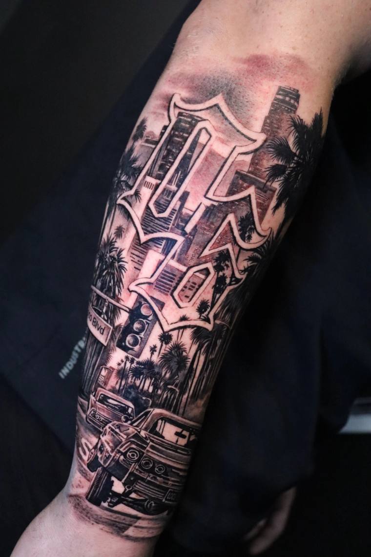 Blackwork forearm tattoo featuring semi-trucks, palm trees, and a cityscape set inside a large Gothic letter M with industria