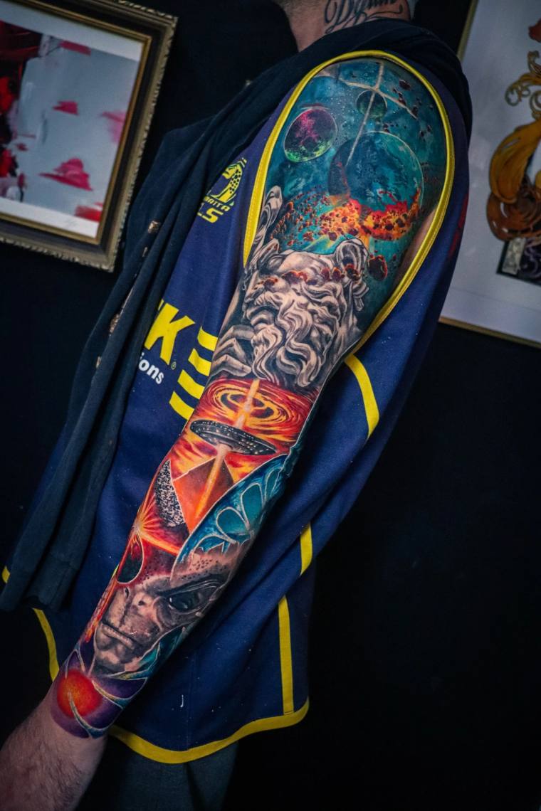 Vibrant full sleeve tattoo featuring a classical sculpted face, cosmic planets, mystical eye, and alien on a deep blue galact