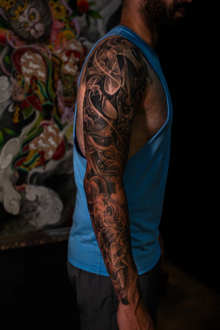 Finished irezumi full sleeve on a man's left arm featuring a hannya mask, lotus flower, and chrysanthemum in black and grey.