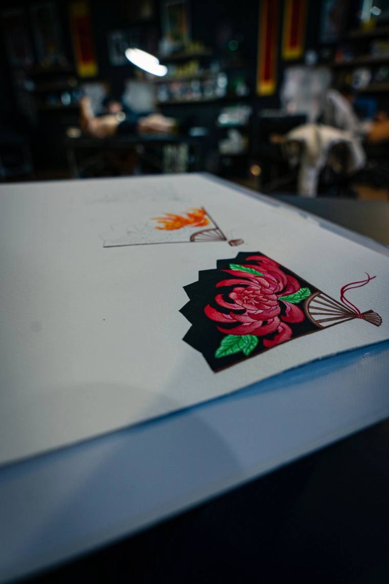 Oriental fan flash, a crimson chrysanthemum blooms across a black folding fan, drawn in watercolour alongside a second design in progress. by Mandy