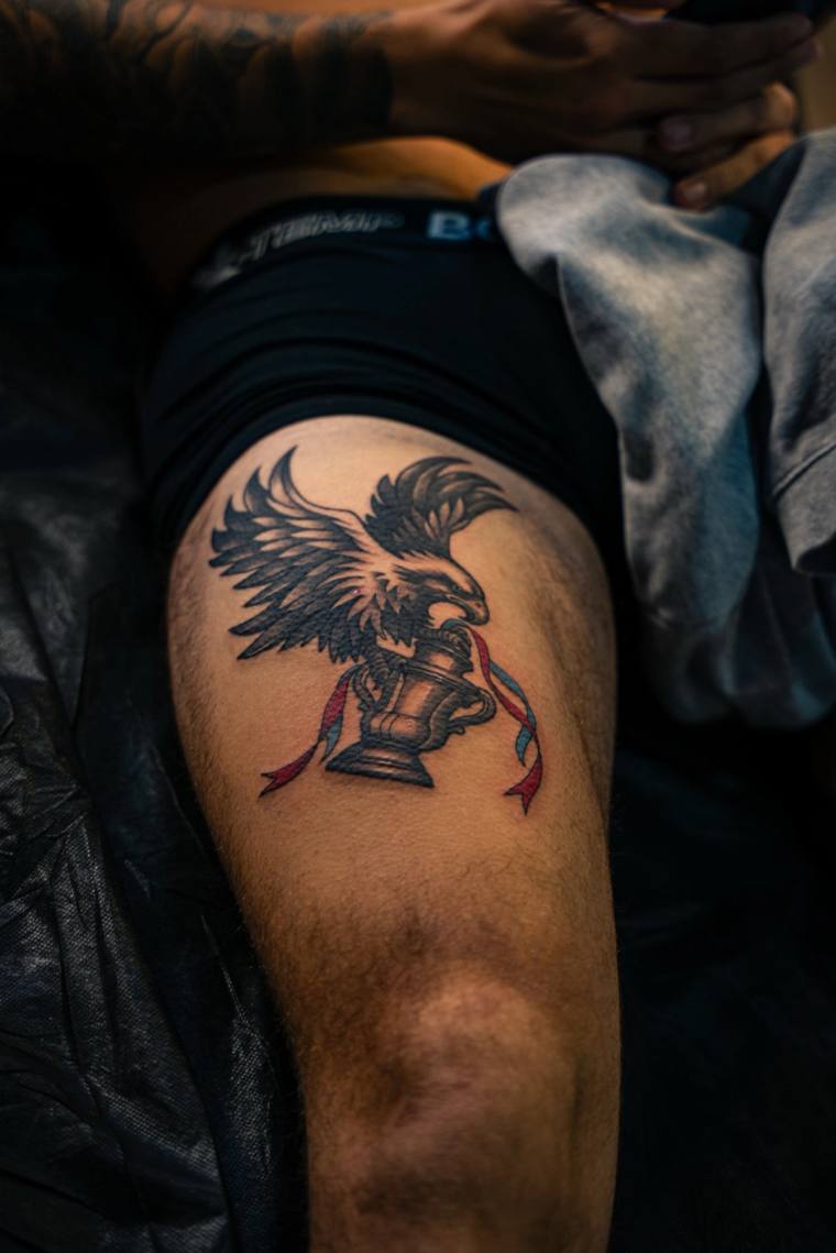 Neo-traditional tattoo on the upper thigh depicting a black eagle clutching a chess rook with red and blue flowing ribbons.