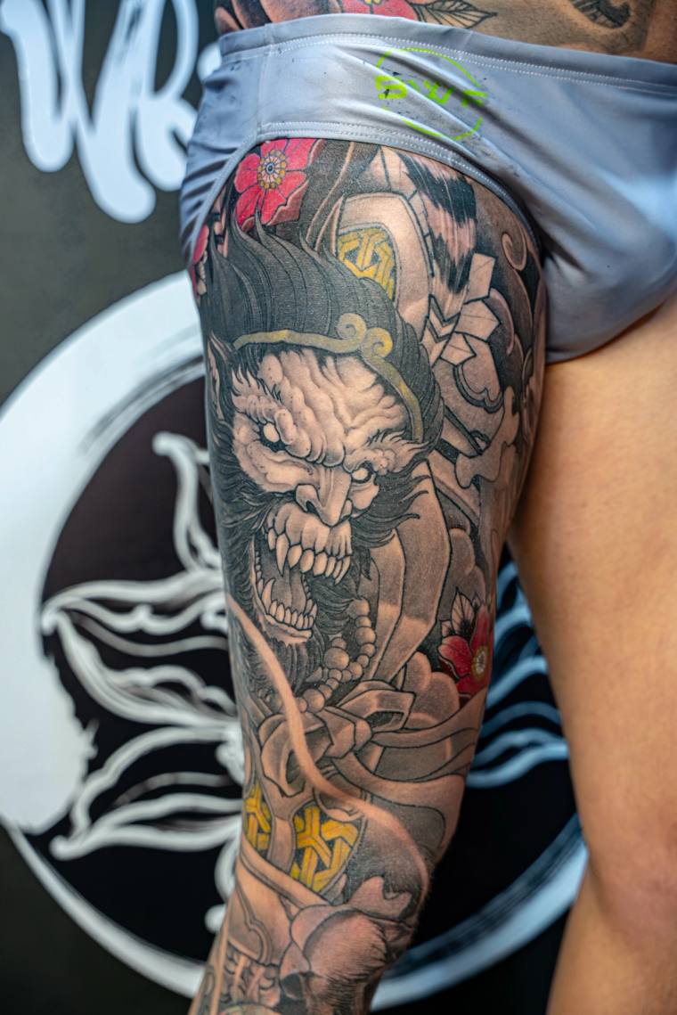 Color irezumi thigh tattoo featuring a snarling fanged demon face and a robed figure accented with red cherry blossoms.