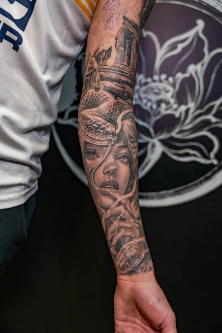 Black and grey forearm tattoo featuring a detailed woman's face intertwined with a snake and architectural elements.