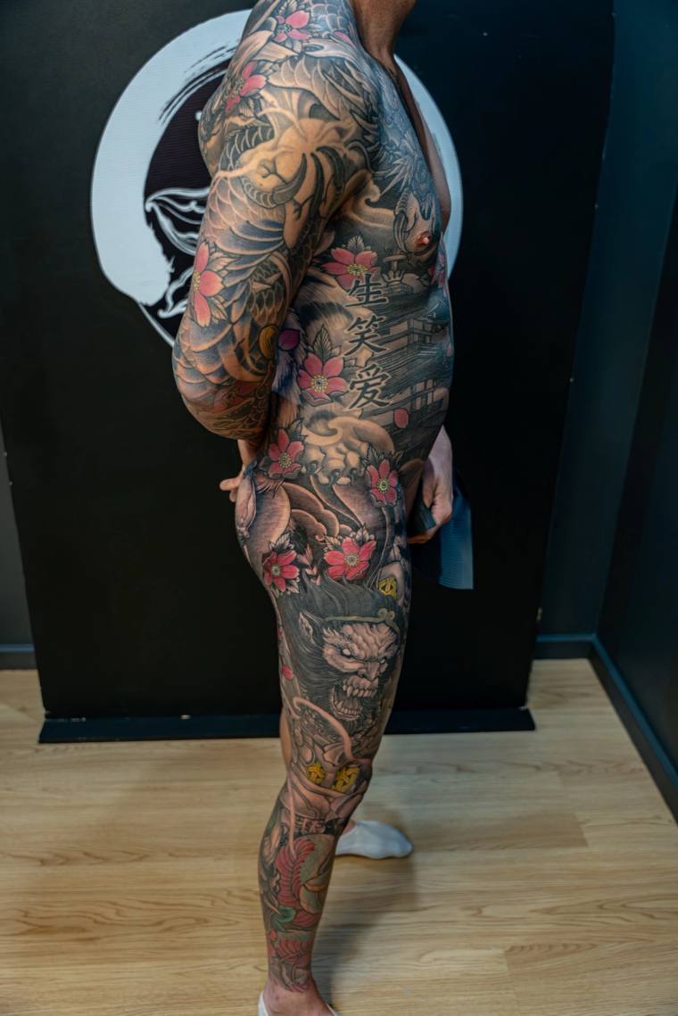 Side profile of a tattooed torso and leg featuring a traditional Japanese irezumi sleeve with koi, hannya mask, and cherry bl