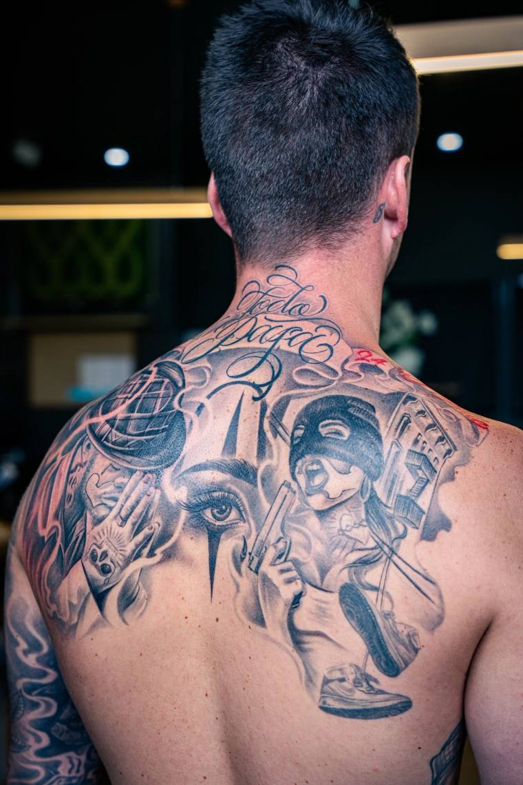 Man displaying a large black and grey back tattoo featuring a masked gunman, streetlamp, weeping eye, playing cards, and scri