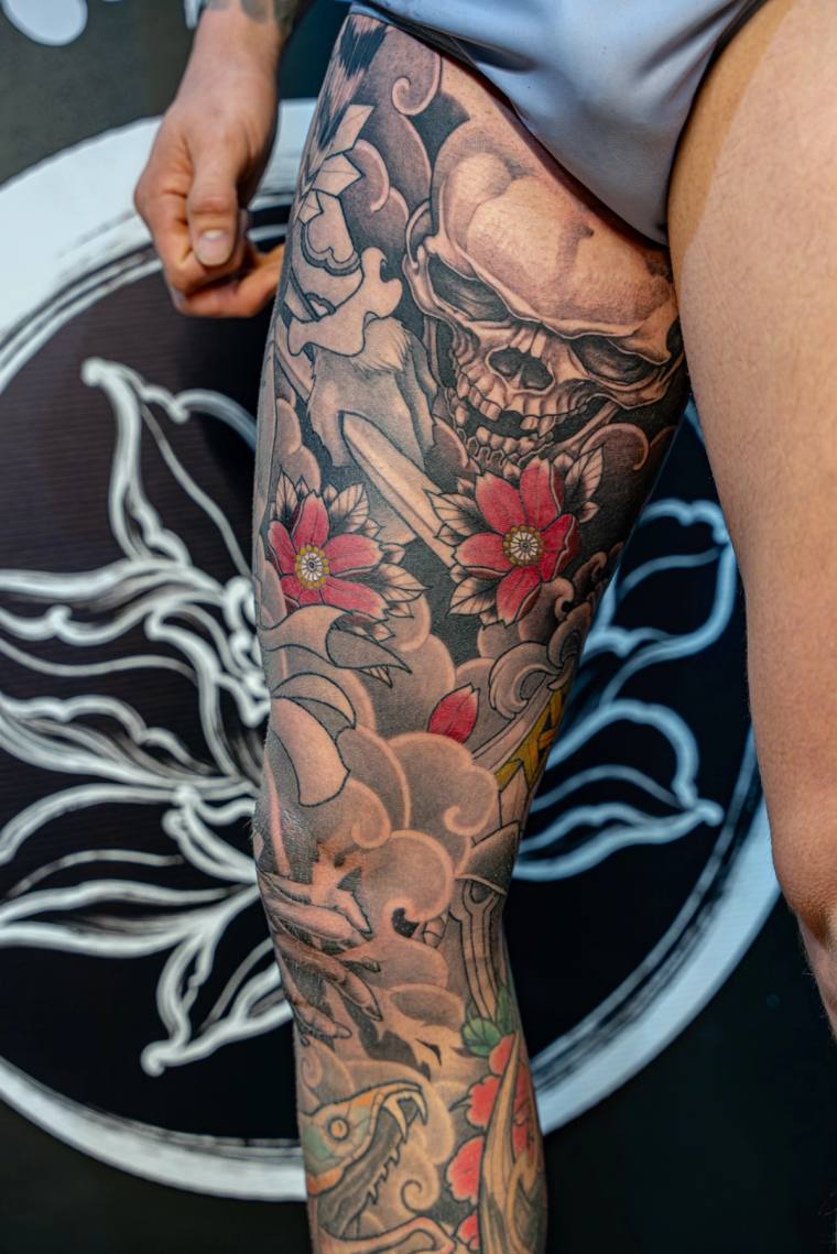 Japanese-style calf tattoo featuring a skull, red peonies, wind bars, and a dragon head in black, grey, and red ink.