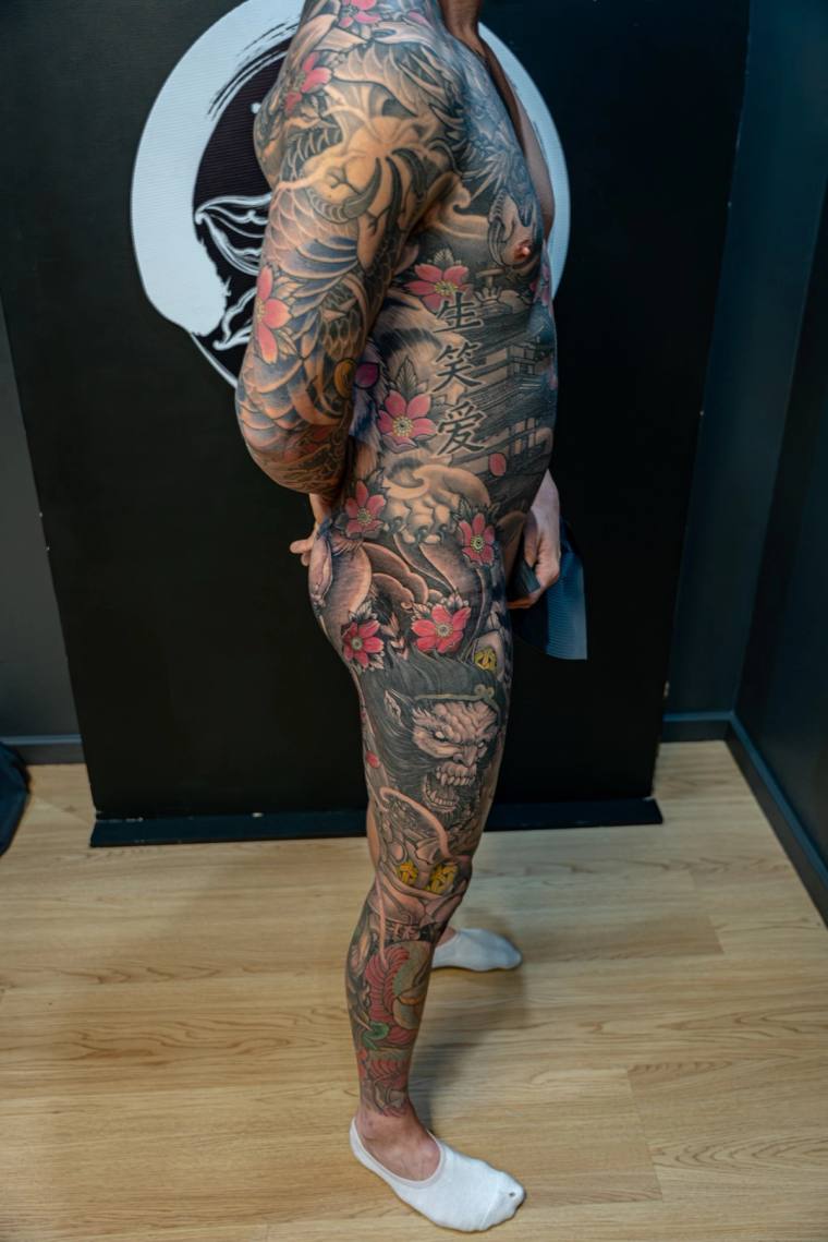 Side view of a person displaying a full irezumi leg and arm sleeve featuring koi, peonies, a hannya mask, clouds, and Japanes