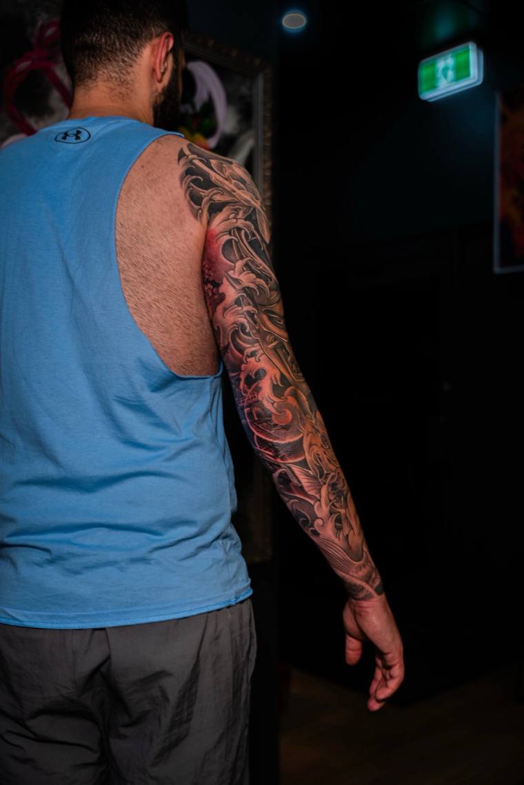 Back view of a man in a blue tank top displaying a finished irezumi full sleeve tattoo with koi, waves, and floral motifs on 