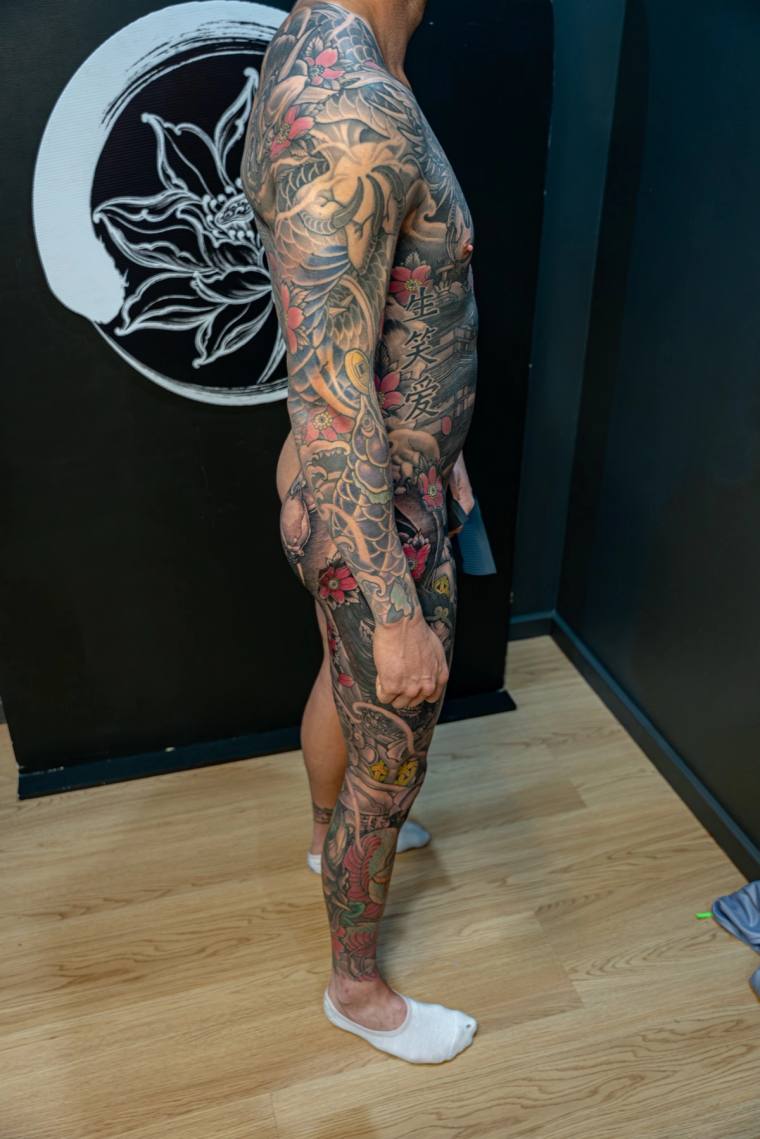 Side profile of a person displaying a full irezumi sleeve and leg piece featuring a tiger, koi, cherry blossoms, kanji, and a