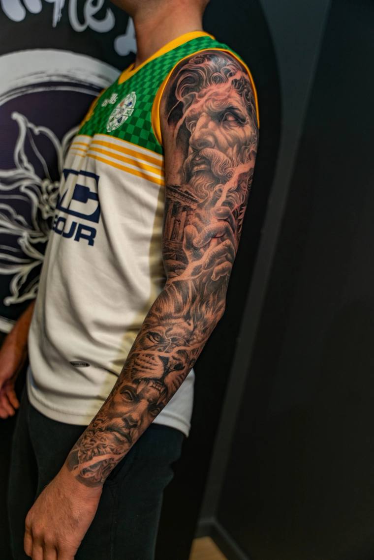 Finished black and grey full-sleeve tattoo featuring classical Greek mythology: a bearded god's face, muscular figures, and s