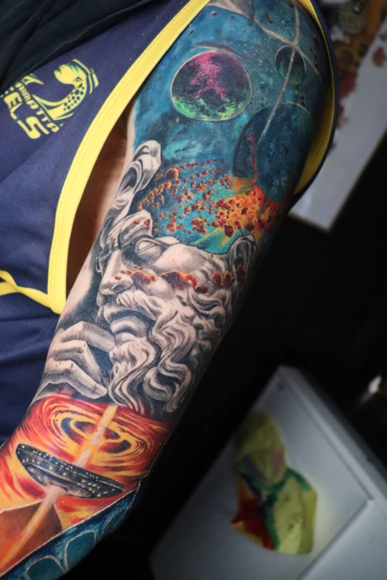 Colorful cosmic sleeve tattoo on upper arm featuring a bearded Greek-style god, planets, stars, a fiery vortex, and a UFO.